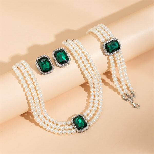 Large green emerald stones adorn this multilayer pearl jewellery set by Eclat New York, which includes a pearl bracelet, matching earrings, and a choker.