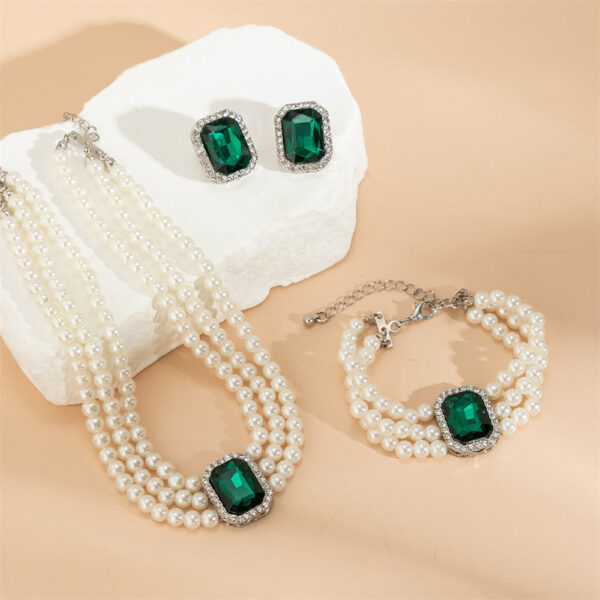 Large green emerald stones adorn this multilayer pearl jewellery set by Eclat New York, which includes a pearl bracelet, matching earrings, and a choker.